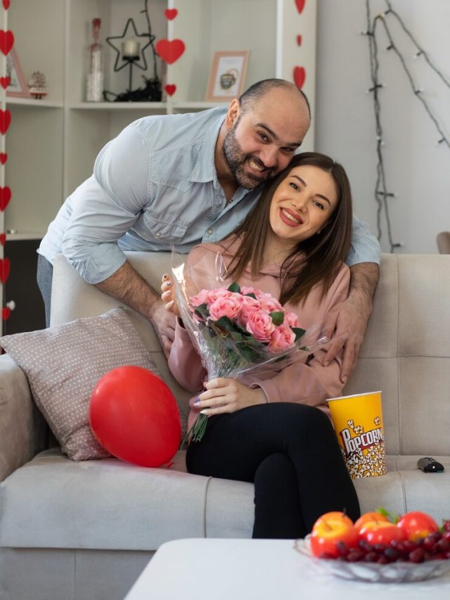jovem-casal-lindo-feliz-dando-um-buque-de-flores-para-sua-namorada-sorridente-sentado-em-um-sofa-em-uma-sala-iluminada-comemorando-o-dia-internacional-da-mulher_141793-61810