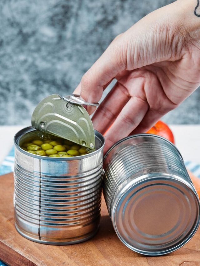 mao-segurando-uma-lata-de-ervilhas-cozidas-em-uma-mesa-branca-com-legumes-e-toalha-de-mesa_114579-24378