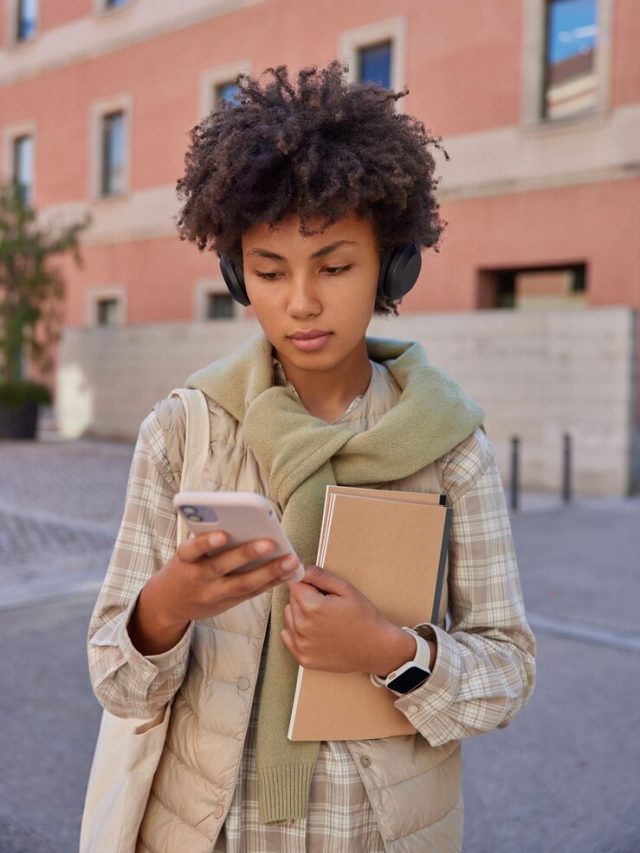 mulher-morena-estudos-on-line-ouve-palestra-educativa-com-smartphone-e-fones-de-ouvido-anda-ao-ar-livre-carrega-blocos-de-notas-vestida-com-roupas-casuais_273609-55644